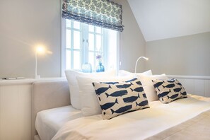 Une chambre à coucher avec un lit, du linge de lit blanc, une fenêtre agrémentée d’une lambrequin à motifs et des lampes de chevet.