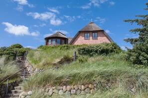 A traditional thatched-roof house with a stone staircase leading up to it, surrounded by lush greenery. - Wattblick. Reet. Sauna. - Wattgeflüster (Sylt)