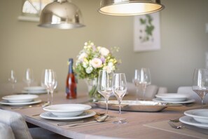 Una mesa de comedor dispuesta con platos blancos, copas de vino y cubiertos, con un jarrón de flores y una botella de vino al fondo.