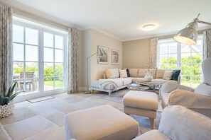 A spacious living room with a large sectional sofa, a coffee table, and a floor lamp. There are large windows providing a view of the outdoors. - Wattblick. Reet. Sauna. - Wattgeflüster (Sylt)