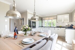 Une cuisine moderne avec des meubles blancs, une table à manger pour quatre personnes et des luminaires suspendus.