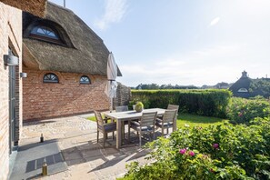 A brick house with a thatched roof, a patio with a table and chairs, and a garden with flowers. - Wattblick. Reet. Sauna. - Wattgeflüster (Sylt)