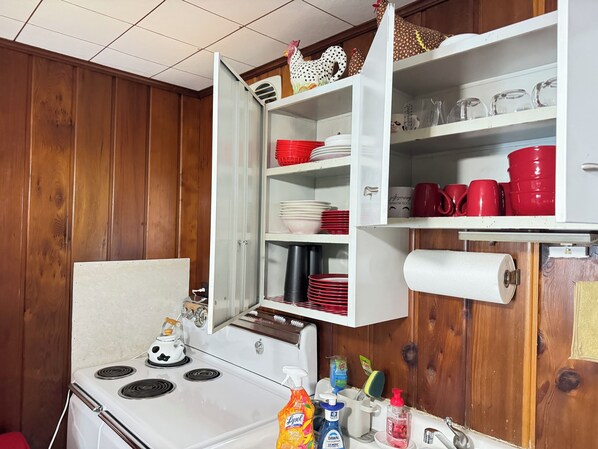 A kitchen with a stove, open cabinets with dishes and utensils, and a wooden wall.