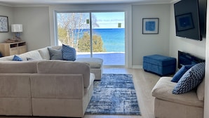 A living room with a large window offering a view of the sea, a sofa, a smaller couch, and a flat-screen TV mounted on the wall.