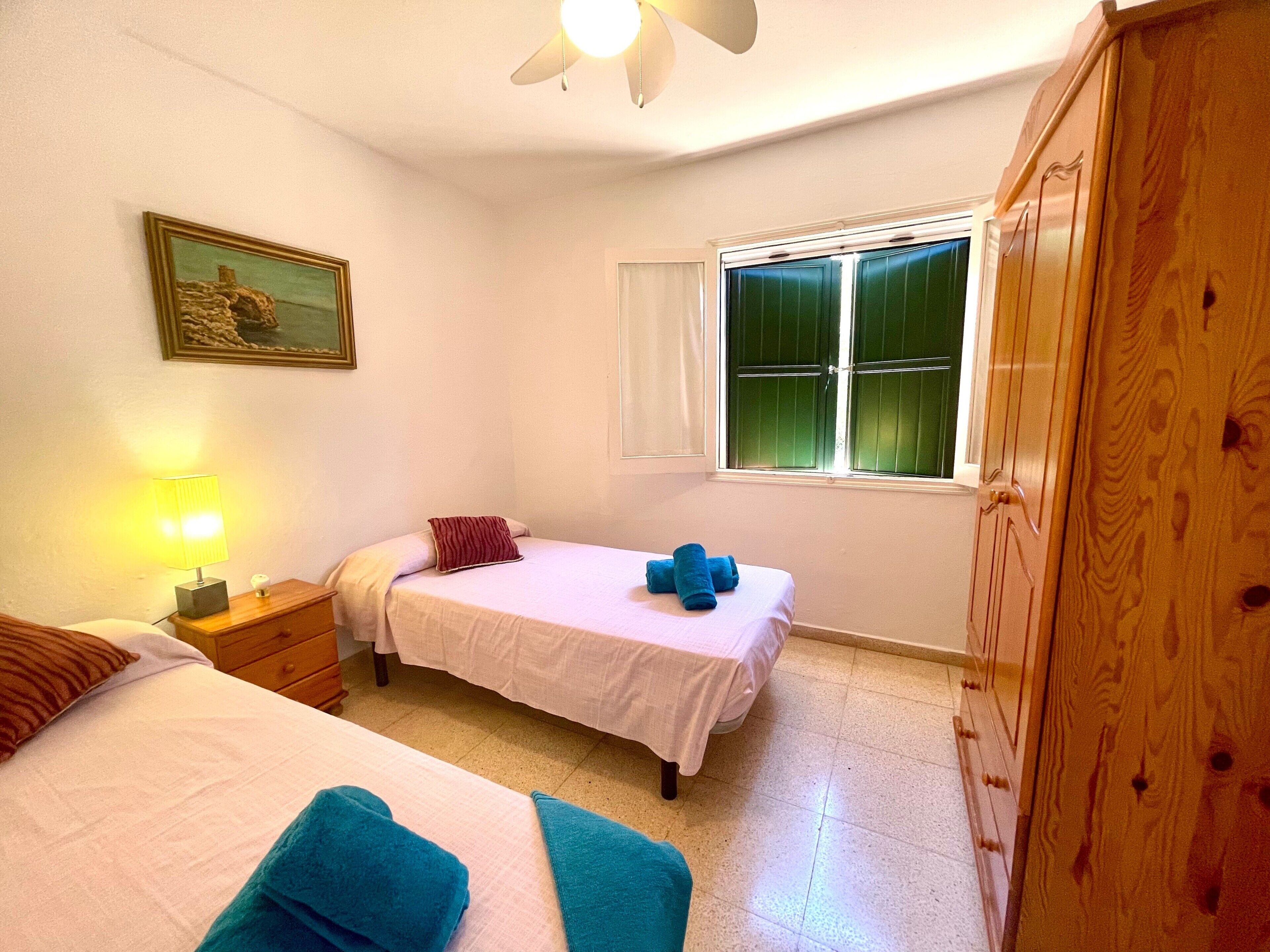 Un dormitorio con dos camas, una mesita de noche con lámpara, una ventana con contraventanas y un armario de madera.