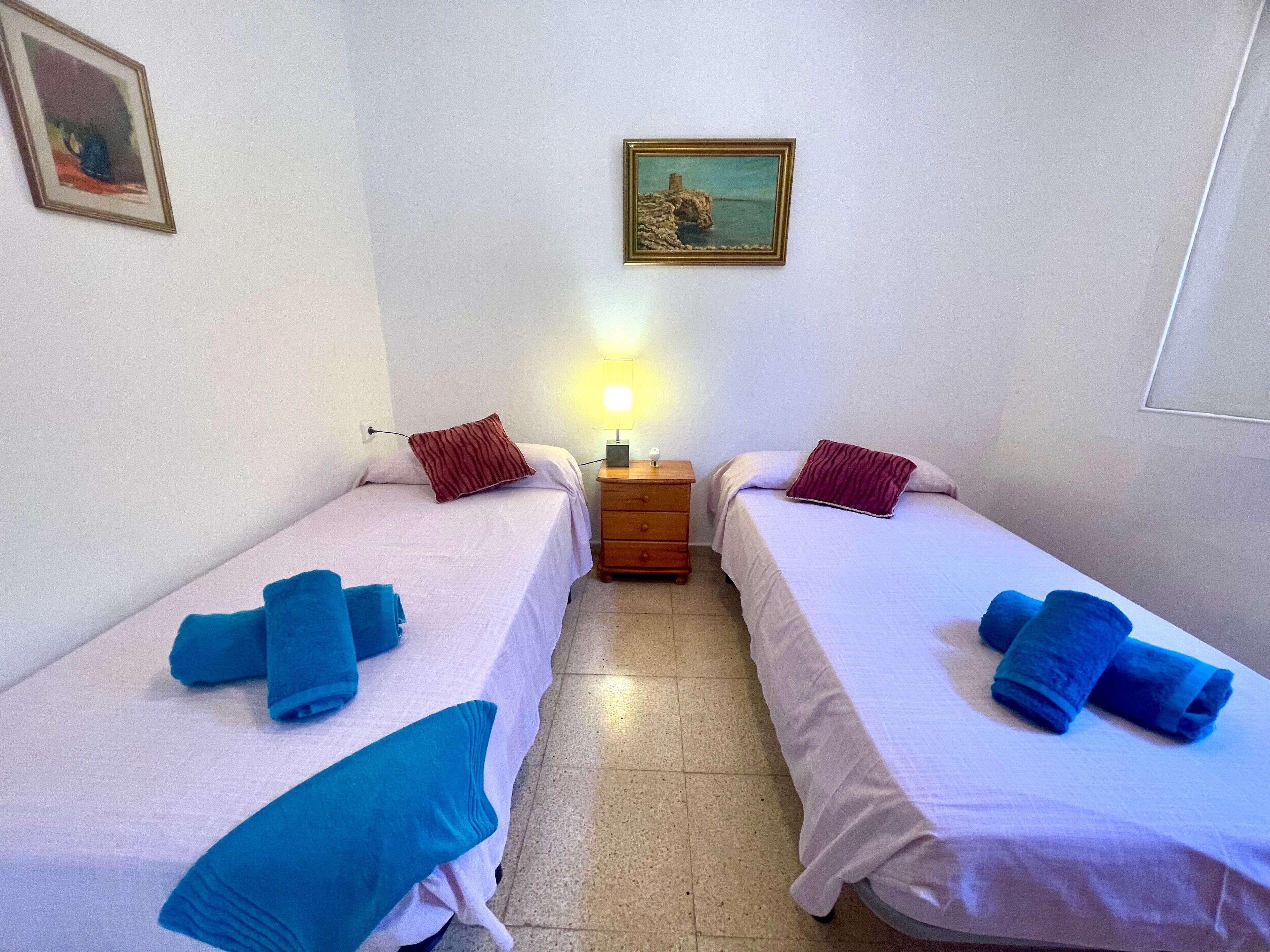 Una habitación con dos camas, cada una con una toalla azul, una mesita de noche de madera con lámpara y un cuadro en la pared.