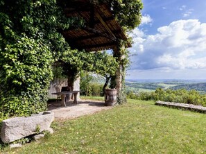 Un área de estar al aire libre de estilo rústico, con un banco de piedra, una mesa de madera y un barril, rodeada de follaje y con vistas a un paisaje pintoresco.