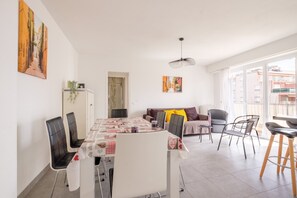 A dining area with a table set for four, a sofa with cushions, and a view of buildings through large windows. - Apartment '2 Rooms Near Beaches Go011' with Balcony and Wi-Fi (Cagnes sur Mer)