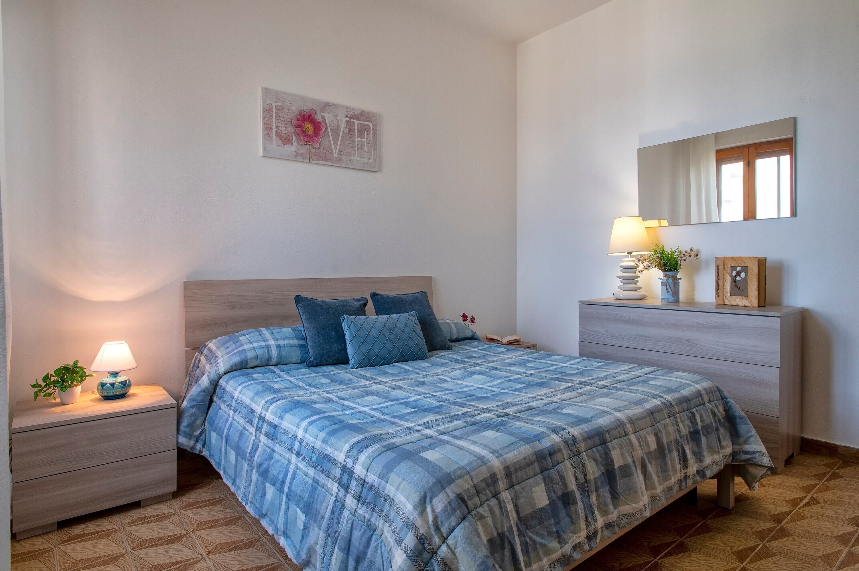 Un dormitorio con cama, mesitas de noche, cómoda, espejo y un adorno de pared.