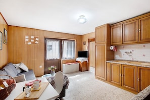 Una habitación compacta con sofá, una mesa de comedor, zona de cocina con armarios de madera, radiador y televisor.