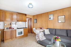 Una cocina compacta con armarios de madera, una estufa blanca y una zona de comedor con un sofá gris y una mesa pequeña.