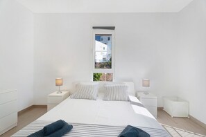 Un dormitorio minimalista con una cama grande, dos mesitas de noche, una ventana con vistas a edificios y un armario blanco pequeño.