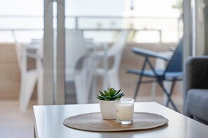 Una mesa con una vela, una pequeña planta en maceta y un vaso de leche, sobre un mantel blanco en una habitación con sillas blancas y vistas a través de un vidrio.
