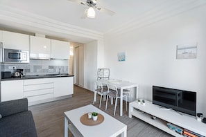 Una cocina moderna con armarios blancos y un microondas, un comedor con una mesa y sillas blancas, un televisor de pantalla plana sobre un soporte blanco y un ventilador de techo.