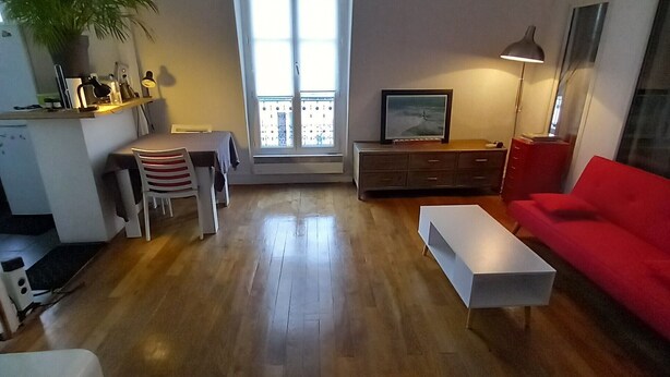 A living room with a wooden floor, a red sofa, a white coffee table, a dining area with chairs, and a large window. - Bright Apartment in Batignolles – Parisian Charm, Self Check-In (Paris)