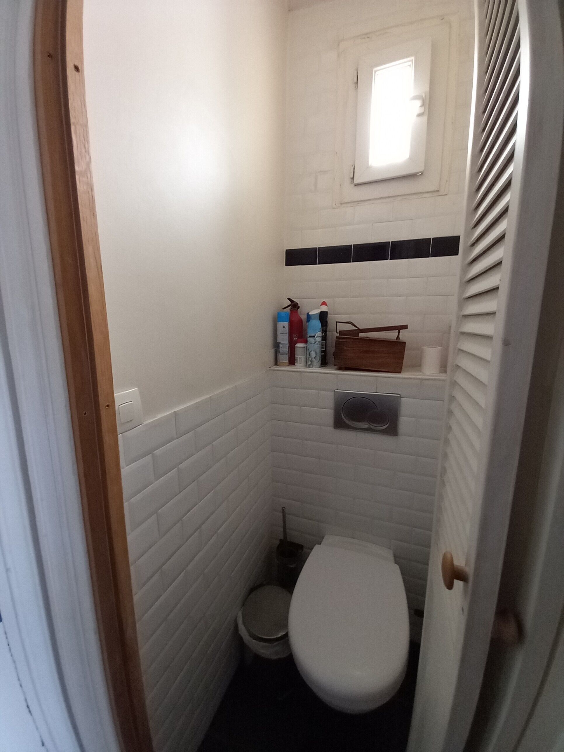 Un bagno piccolo rivestito di piastrelle bianche, con il water, una piccola finestra e uno scaffale con articoli da toilette.