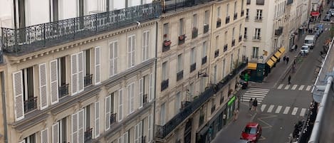 A row of multi-story buildings with balconies, a street with cars and pedestrians, and storefronts.