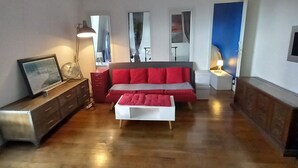A spacious living room with a wooden floor, a red sofa, a white ottoman, a sideboard, and a mirror. - Bright Apartment in Batignolles – Parisian Charm, Self Check-In (Paris)