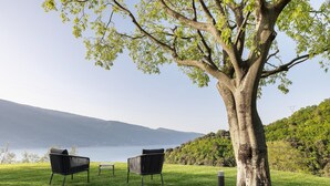 Eine begrünte Außenanlage mit zwei schwarzen Stühlen und einem Tisch, einem großen Baum und Blick auf einen See und Berge.