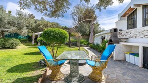 An outdoor patio with a glass table and wicker chairs, surrounded by greenery and a stone pathway.
