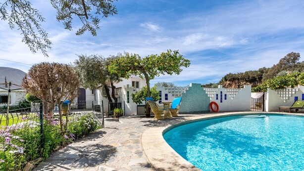 A swimming pool with stone tiles, a white building with blue windows, and a tree in the foreground.