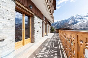 Un bâtiment en pierre doté de grandes fenêtres, d’un balcon en bois et surplombant un paysage montagneux.
