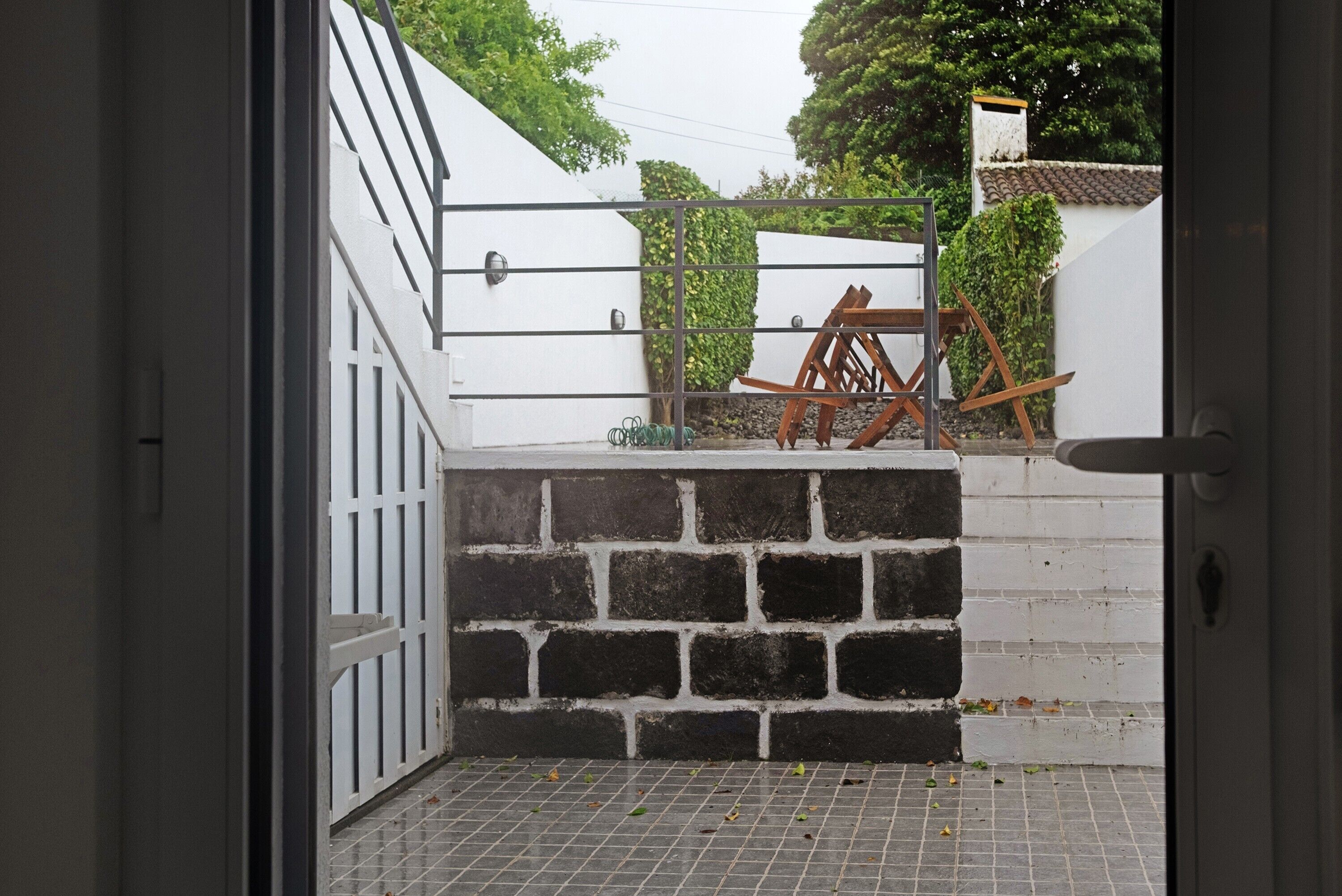On aperçoit, à travers une porte ouverte, une cour intérieure avec un mur de briques, une porte en métal et une chaise en bois.