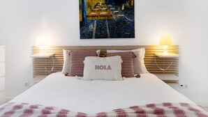 Een bed met een witte deken, twee bedlampen, een schilderij boven het bed, en een kussen met 'HOLA' erop.
