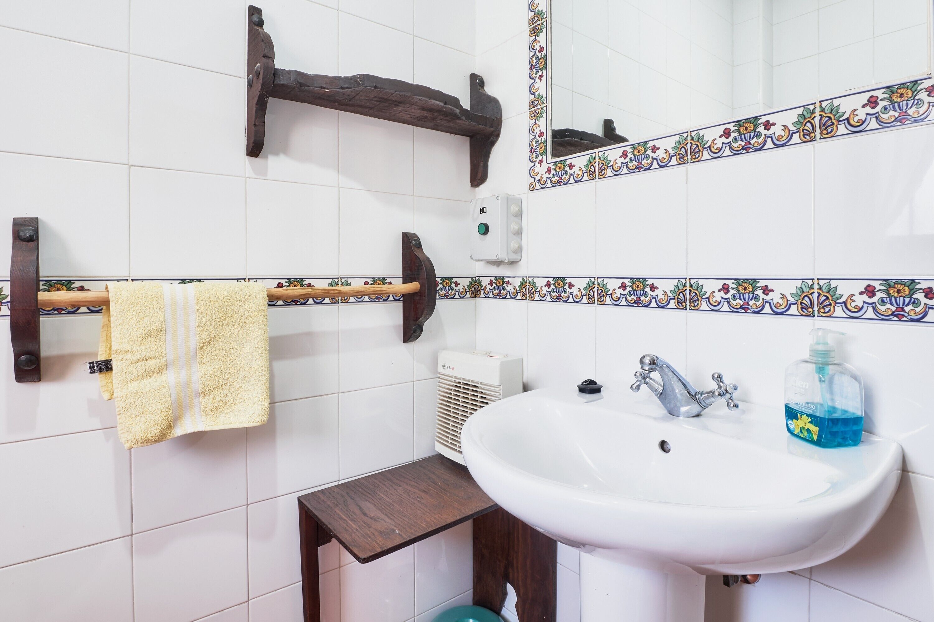 A bathroom with a white sink, a mirror with decorative tiles, a towel rack with a yellow towel, and a wooden shelf.