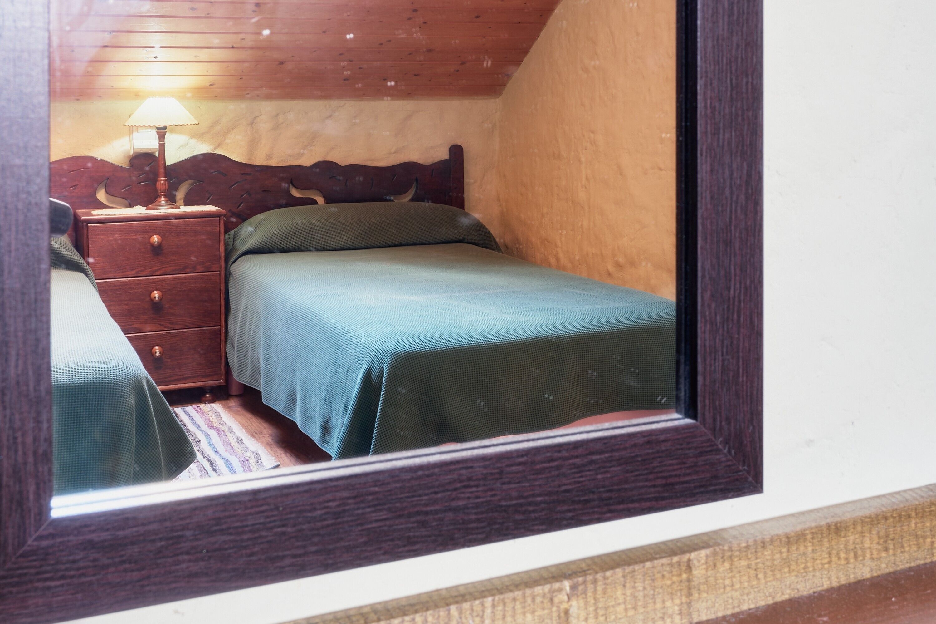 A bedroom with a wooden headboard, a single bed with a green bedspread, a nightstand with a lamp, and a small drawer.