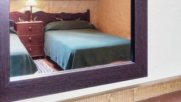 A bedroom with a wooden headboard, a single bed with a green bedspread, a nightstand with a lamp, and a small drawer.