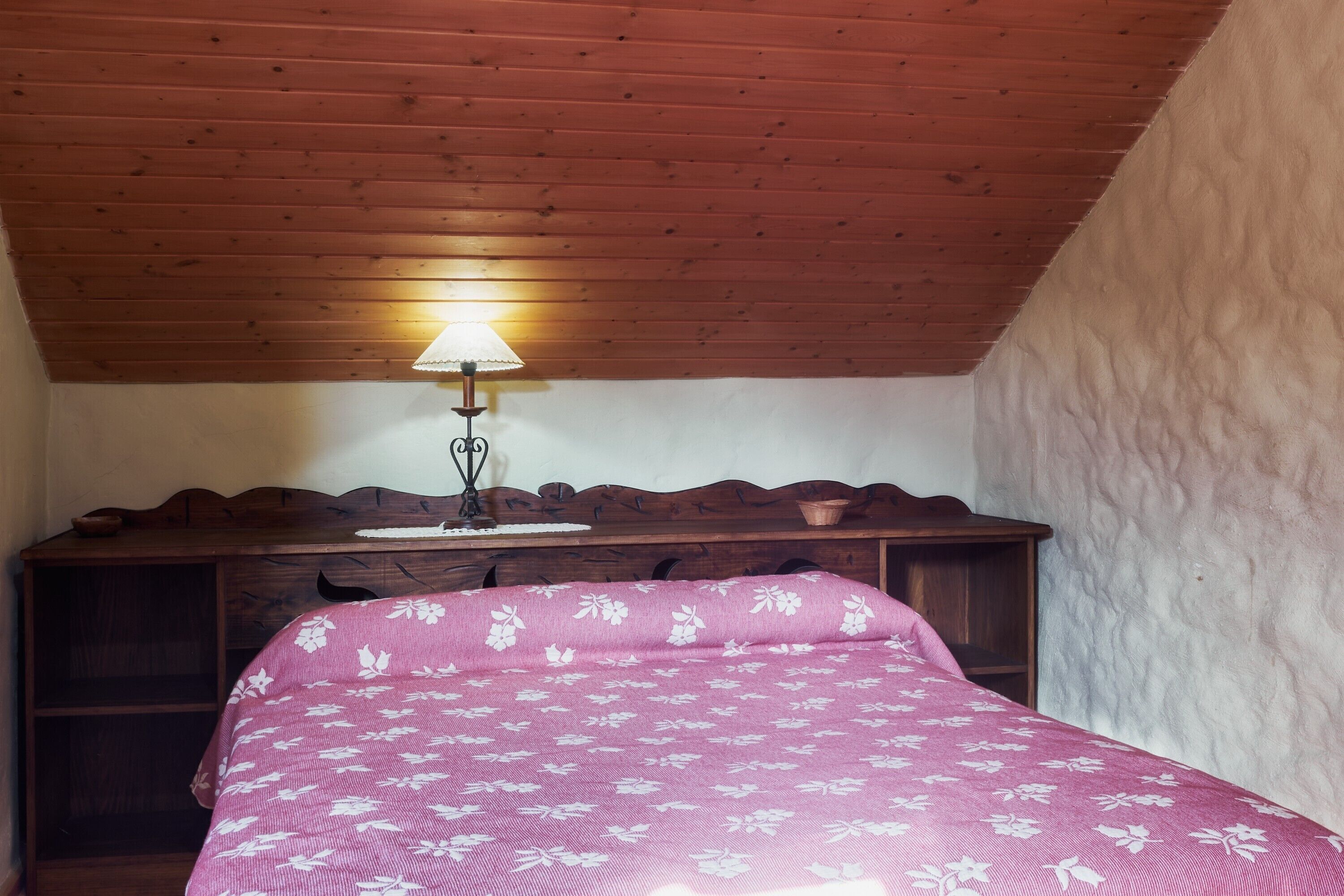 A bedroom with a wooden ceiling, a bed with a floral bedspread, a nightstand, and a lamp.