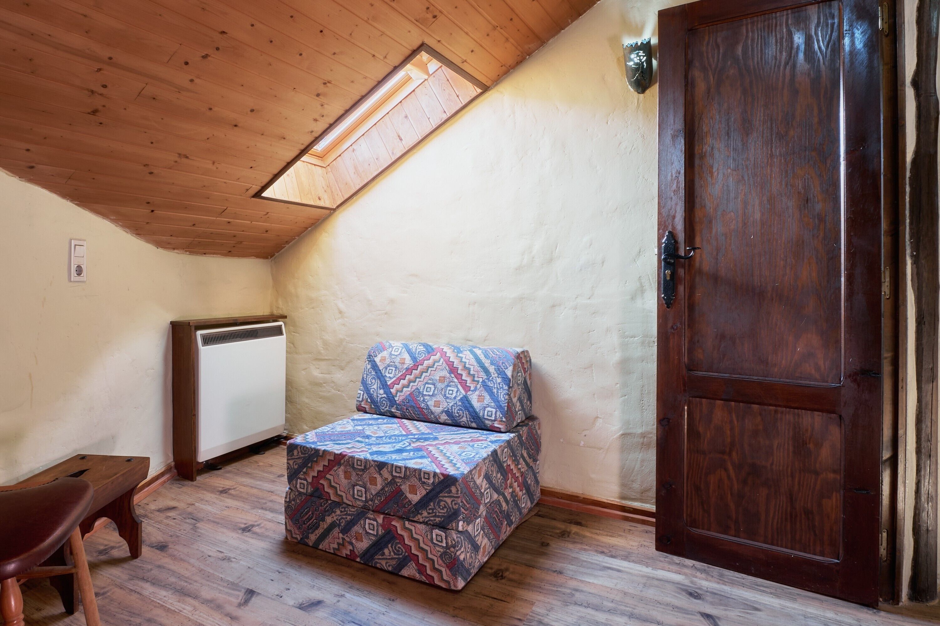 Une pièce avec un plafond en pente, un banc en bois, un canapé à motifs, une porte et un radiateur.
