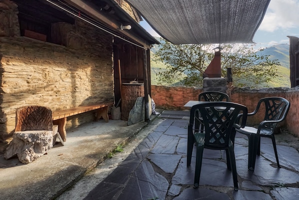 A stone patio with a wooden bench and plastic chairs, a stone wall, and a view of mountains.