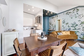 A dining area with a wooden table and chairs, a kitchen in the background, and a living area with a sofa and floral wallpaper.