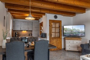 A cozy kitchen with wooden beams, a dining area with grey upholstered chairs, and a television displaying a marina scene. - 50005 Ferienhaus Louis - Die Doppelhaushälfte mit ca. 78qm Kann mit bis zu 6 Personen und Einem (Wittmund)