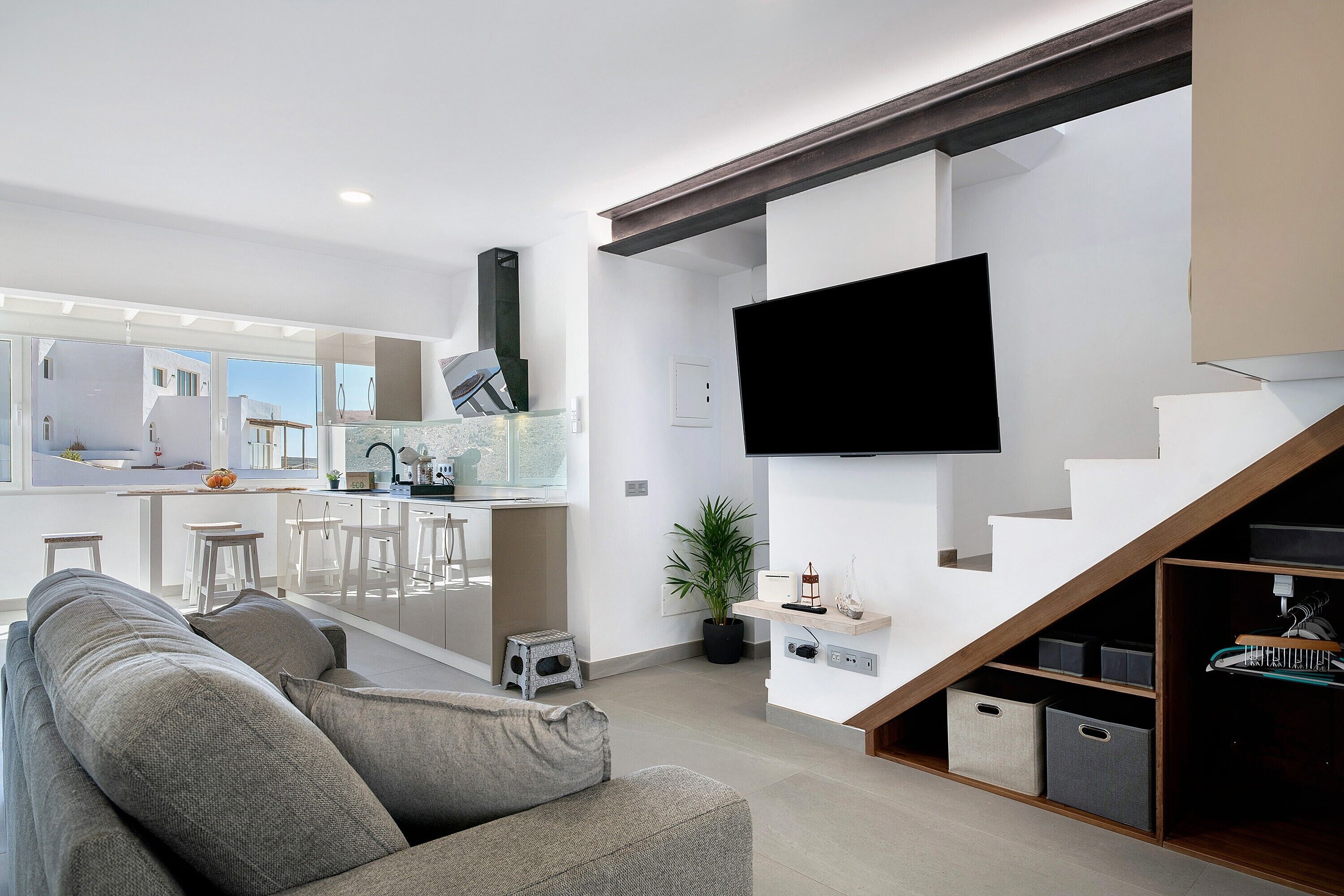 A modern living room with a sofa, a TV mounted on the wall, a kitchen in the background, and a staircase with storage.