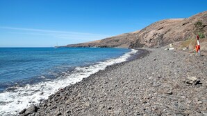 Una playa rocosa con cielo azul despejado y un velero a lo lejos.