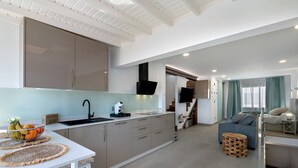 Una cocina moderna con encimera blanca, horno integrado y lavamanos. Se observa una zona de comedor con mesa y taburetes blancos. Al fondo, se distingue una sala de estar con sofá y mesita de centro.