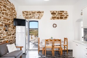 Una cocina con armarios blancos, una zona de comedor con sillas de madera y una mesa, un muro de piedra y vistas a través de una puerta abierta.