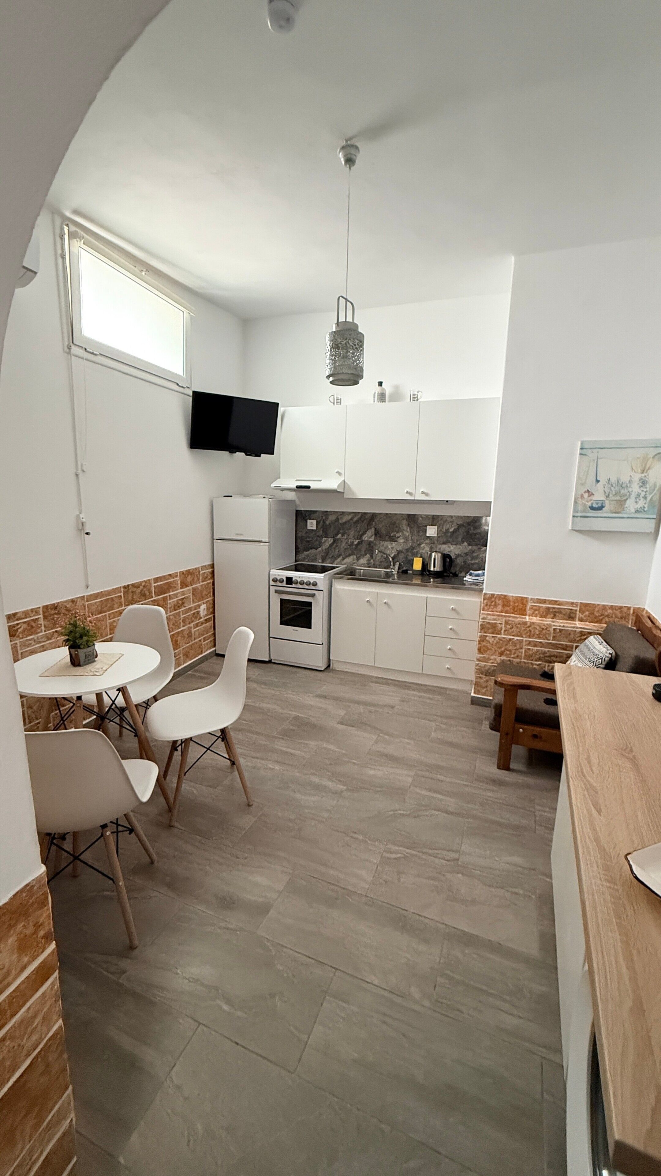 A compact kitchen with a built-in oven, a small dining area with chairs, a TV mounted on the wall, and a stone backsplash.
