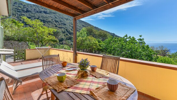 Une table en bois recouverte d’une nappe à carreaux, des chaises et un vase rempli de fleurs, le tout sur un balcon donnant sur un paysage montagneux.