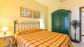 Une chambre à coucher avec un lit, deux lampes fixées au mur, une armoire verte et un ventilateur sur pied.