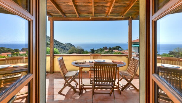 Een overdekt terras met een tafel en stoelen, met uitzicht op een prachtig uitzicht over zee en heuvels.