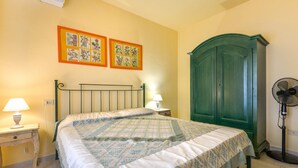 Een slaapkamer met een bed, nachtkastje, lamp, wanddecoratie, kledingkast en een ventilator.