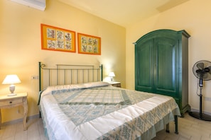 A bedroom with a bed, nightstand, lamp, wall art, wardrobe, and standing fan.