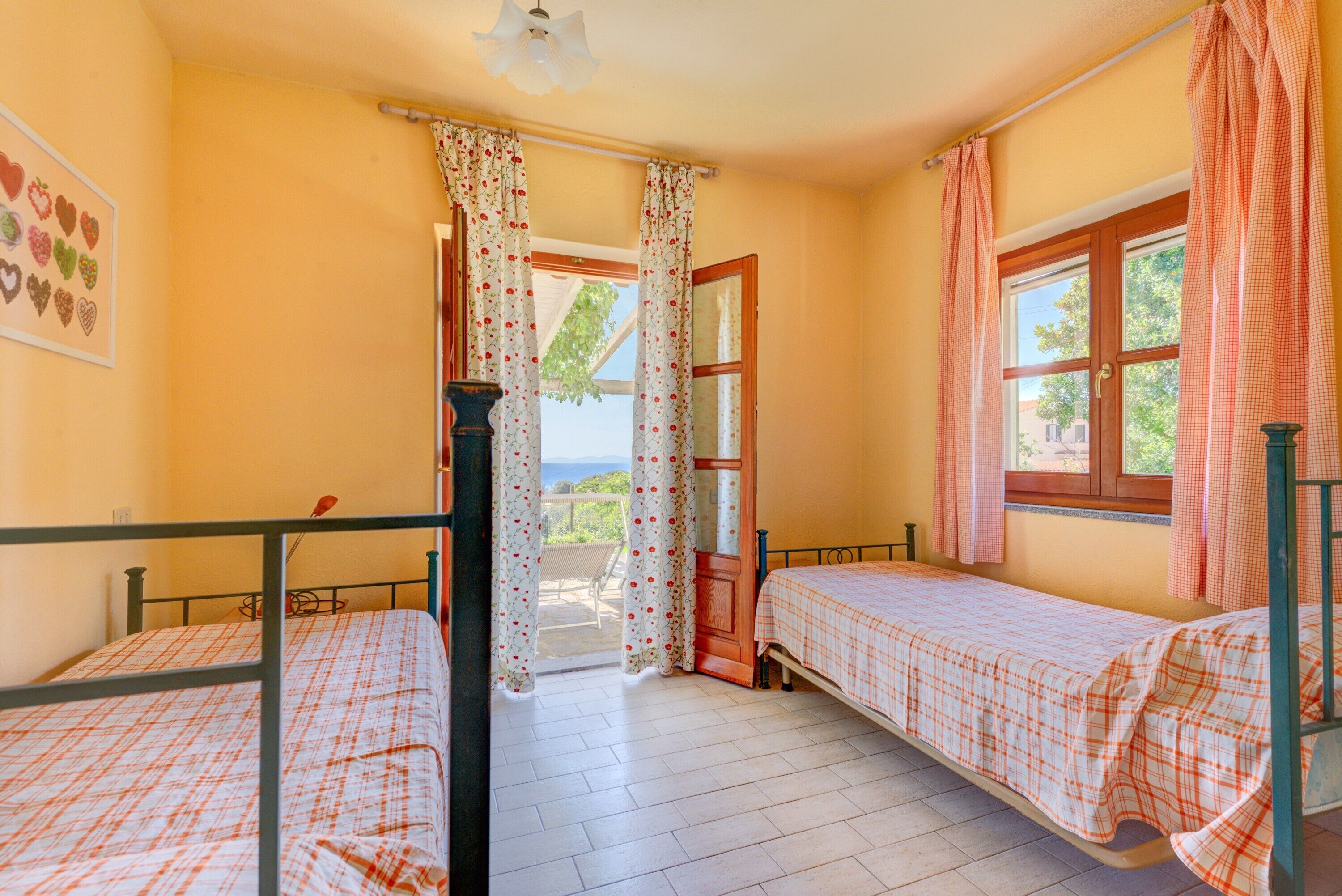 A room with two beds, a window with curtains, a door leading outside, and a view of the outdoors.