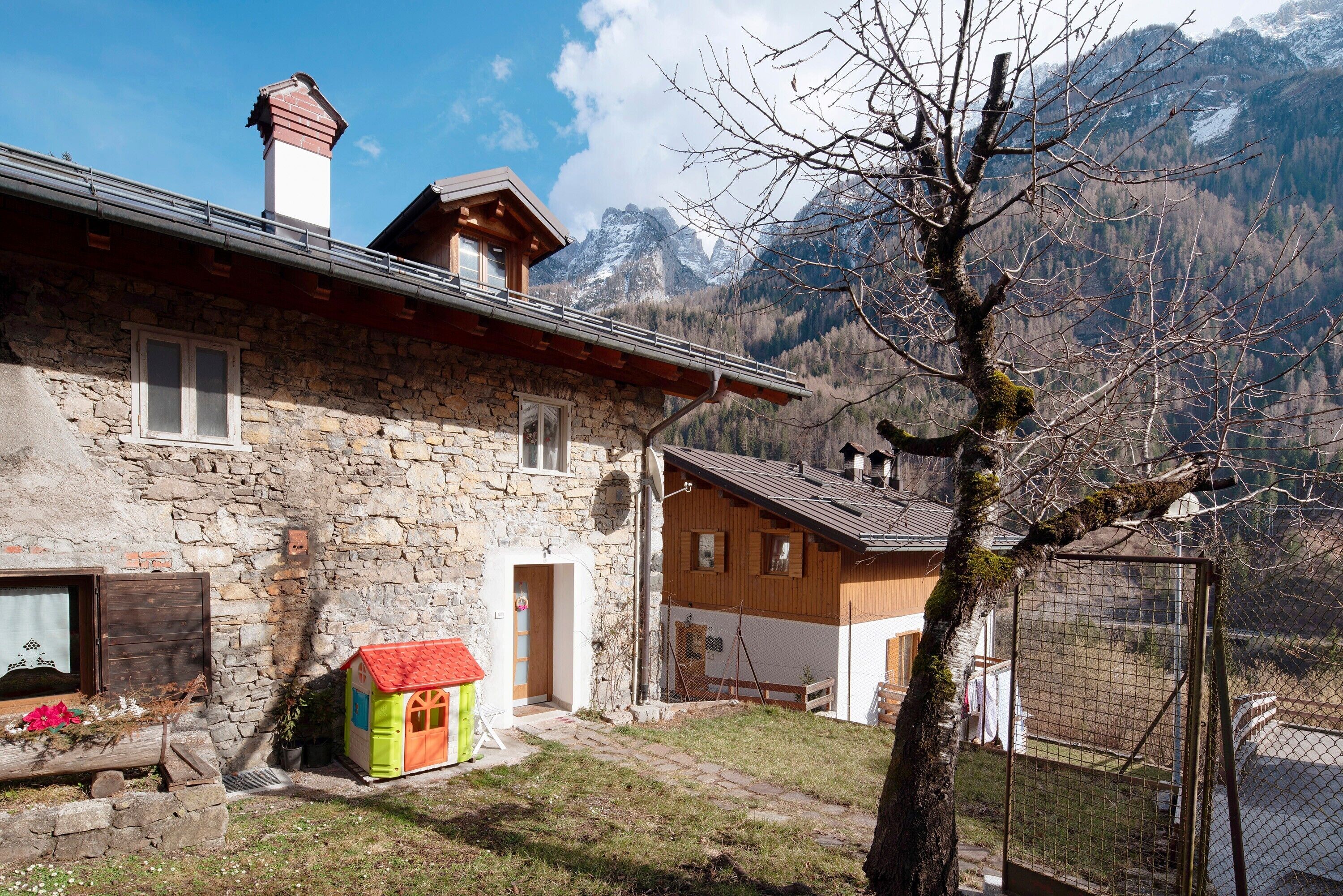 Ein Steinhaus mit Anbau aus Holz, ein kleines Spielhäuschen und ein eingezäunter Bereich mit Bergen im Hintergrund.