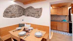 Un comedor con una mesa de madera dispuesta para cuatro personas, detalles en pared de piedra y armarios de madera en la cocina contigua.