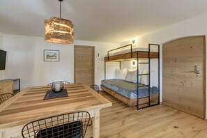 Een kleine kamer met een stapelbed, een houten eettafel met stoelen, een hanglamp en een deur met een houten paneel.